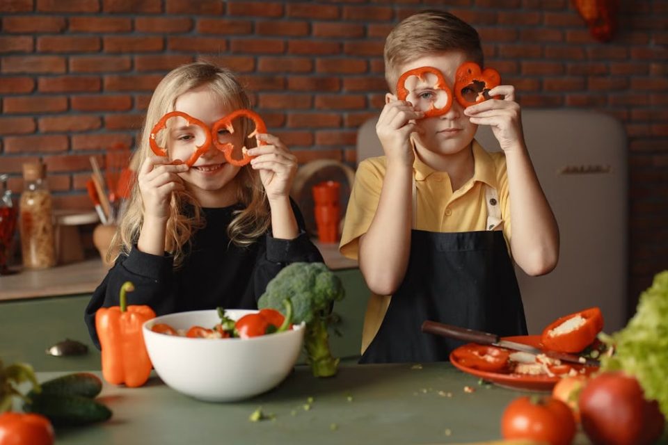 Funny boy and girl wearing apron looking through slices of pepper while preparing healthy vegetarian salad together at table with ingredients and bowls